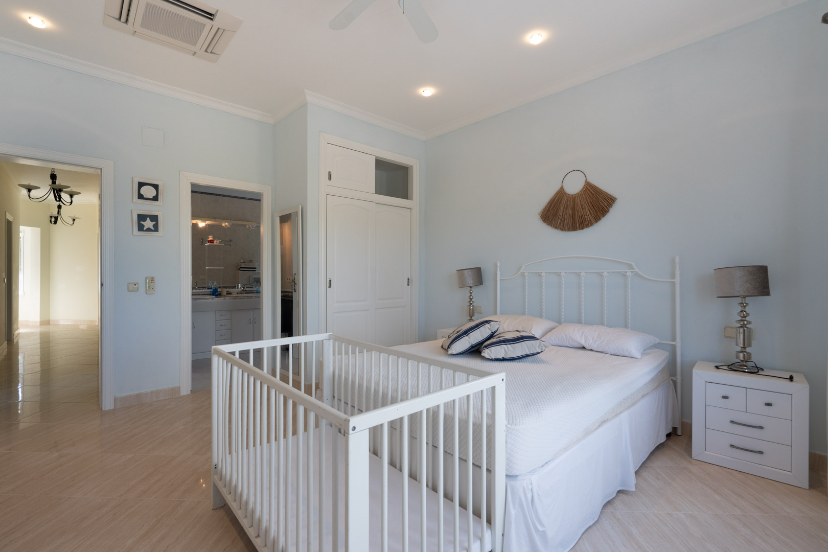 Bright bedroom featuring pale blue walls, a white wrought iron headboard, a wooden crib, built-in wardrobes, and access to an ensuite bathroom.