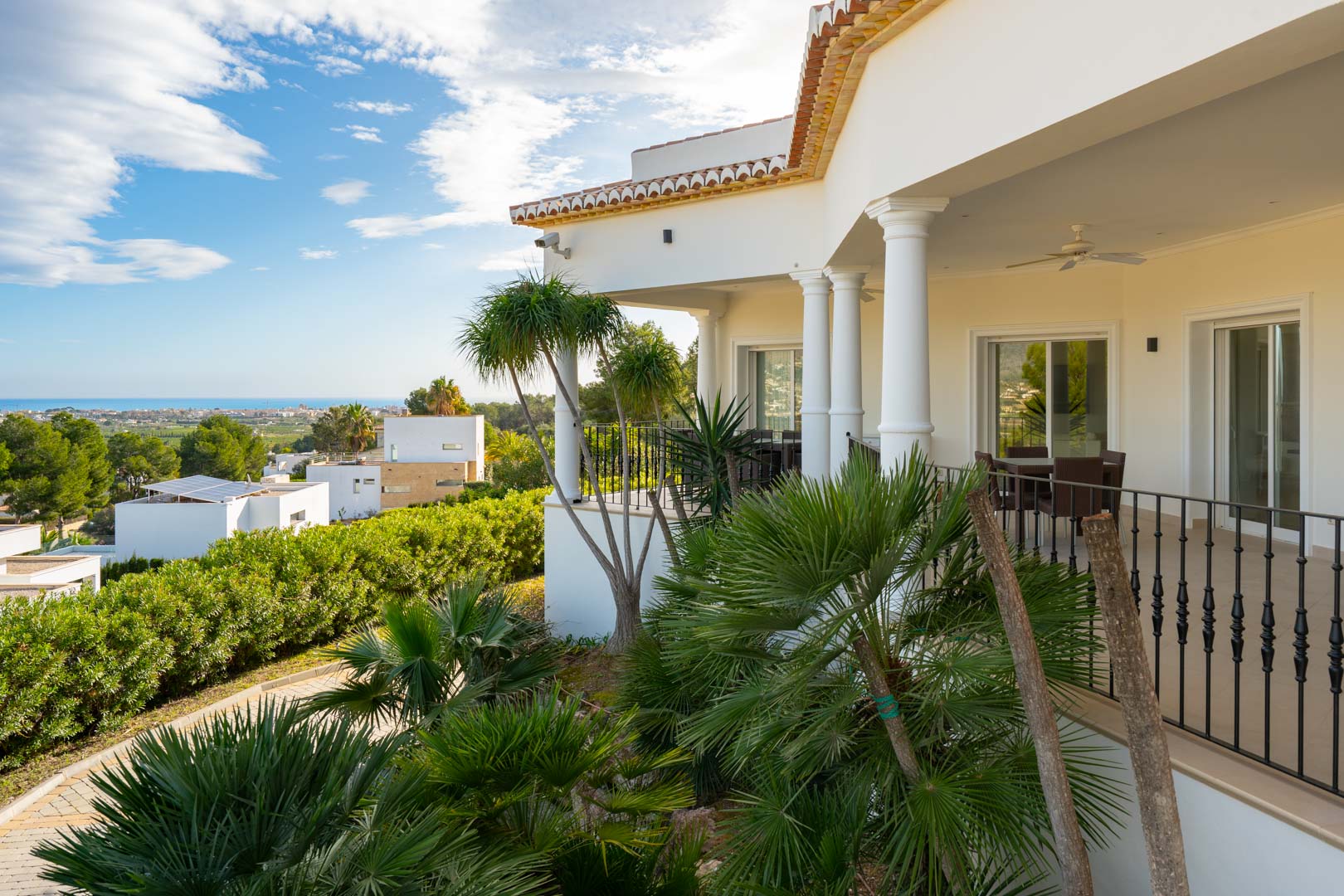 Covered terrace featuring classical columns and wrought iron railings, integrated with tropical palms and offering panoramic views of the Mediterranean coastline.