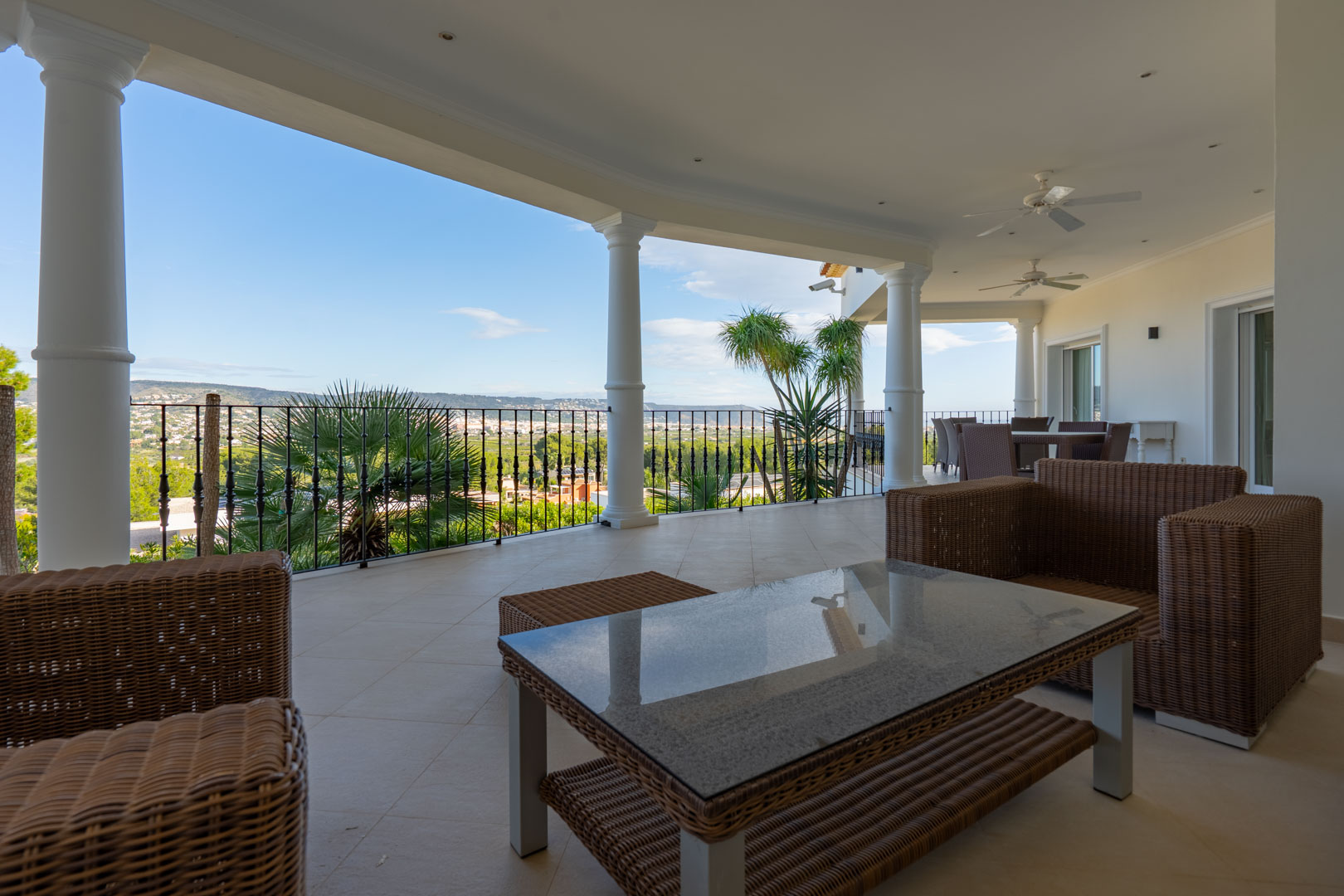 Covered terrace featuring white columns, wicker lounge furniture, and panoramic views of the valley and Montgó. Includes ceiling fans and recessed lighting.
