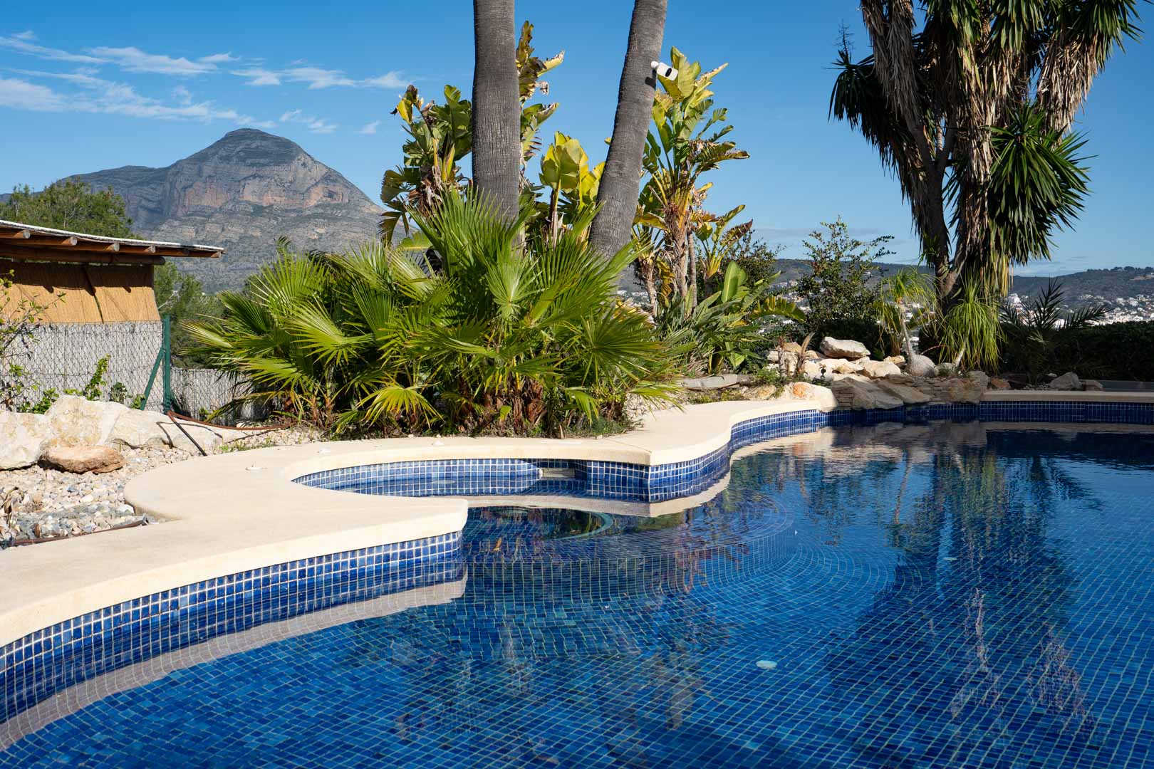 Curved blue mosaic pool featuring an integrated jacuzzi, framed by lush tropical gardens and clear views of the iconic Montgó mountain.