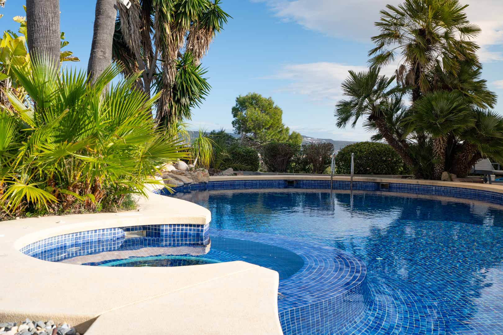 Curved outdoor swimming pool with blue mosaic tiling and integrated steps, surrounded by lush tropical gardens and palm trees.