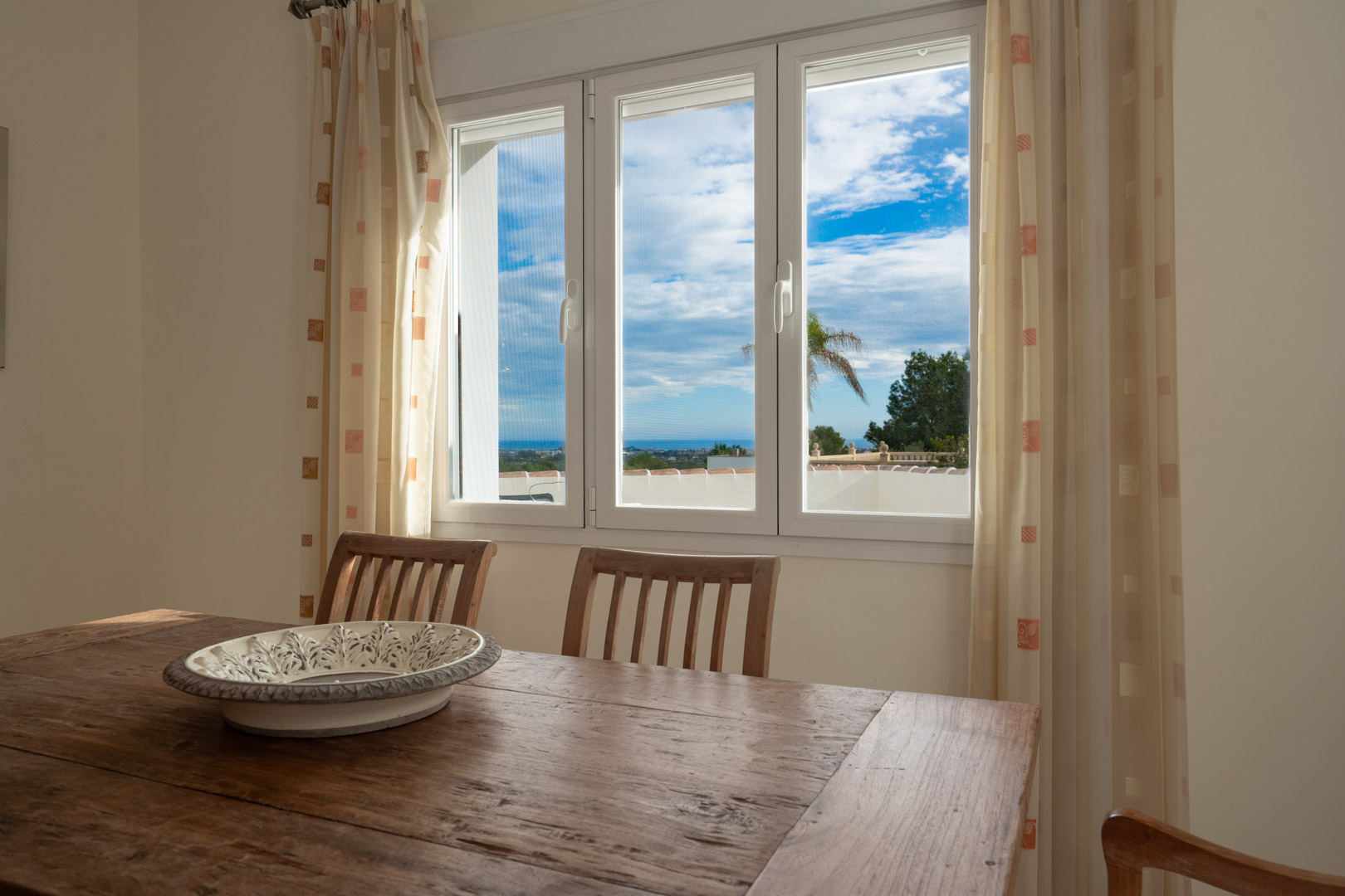 Dining area featuring a rustic wood table and chairs with views of the Mediterranean Sea through a white triple-pane window with natural light.