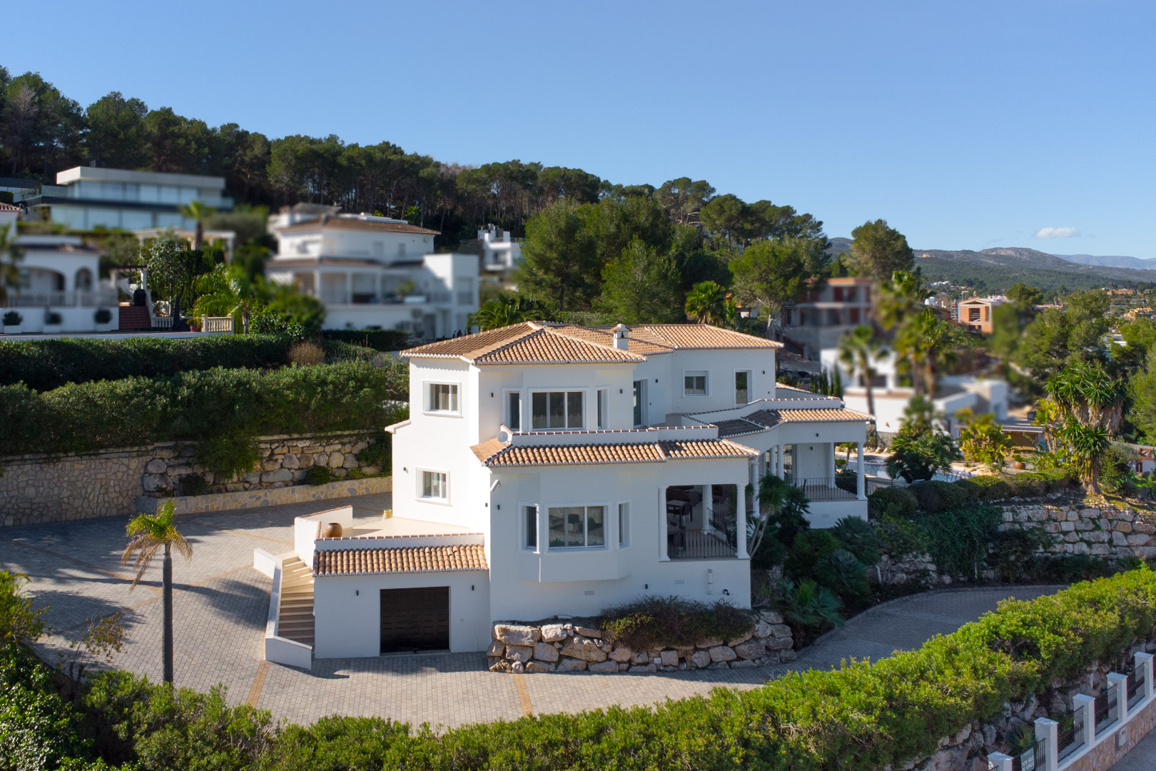 Elevated exterior view of the white Mediterranean villa featuring multi-level terracotta roofs, a spacious paved driveway, and an integrated lower-level garage.