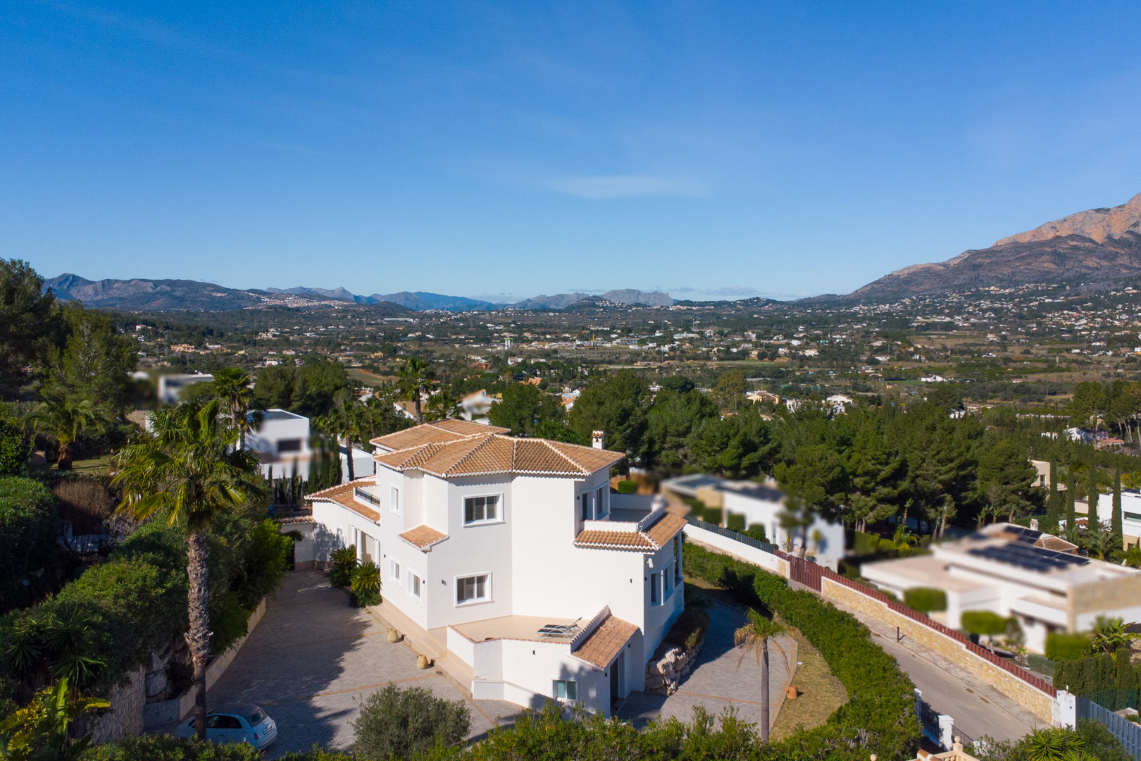 Elevated exterior view of the white villa with terracotta roofs, featuring a spacious paved driveway and panoramic views of the valley and Montgó mountain.