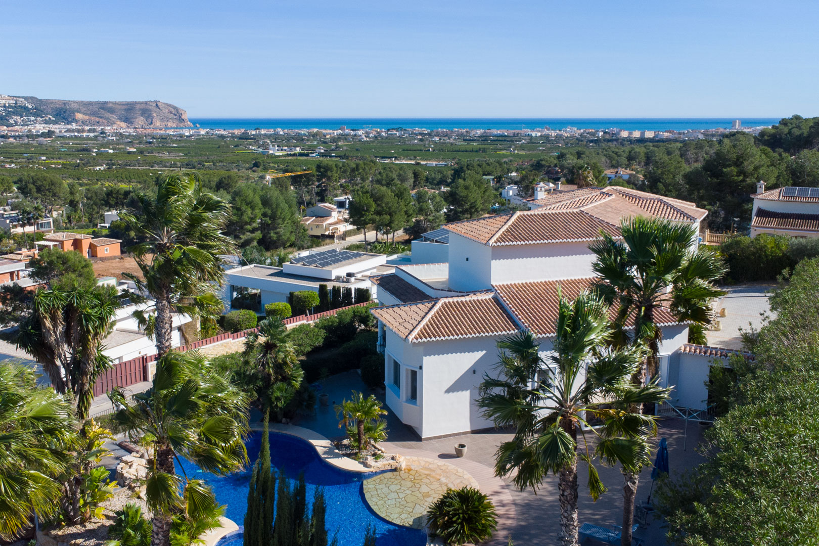 Elevated view of the white villa featuring a blue pool, mature palm trees, and panoramic vistas of the Mediterranean coastline.