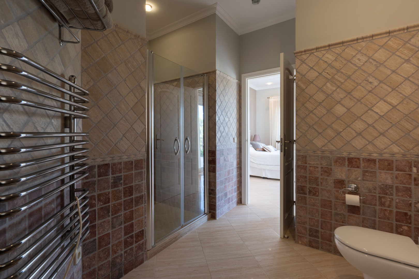 En-suite bathroom featuring a glass-enclosed shower, chrome heated towel rail, and decorative earth-toned tiling with a view into the bedroom.