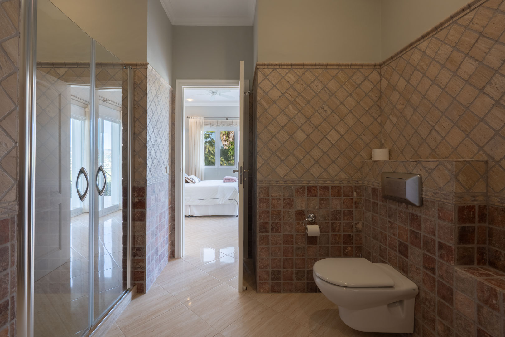 En-suite bathroom featuring rustic stone tiling, a wall-hung toilet, and a doorway leading into the bright master bedroom.