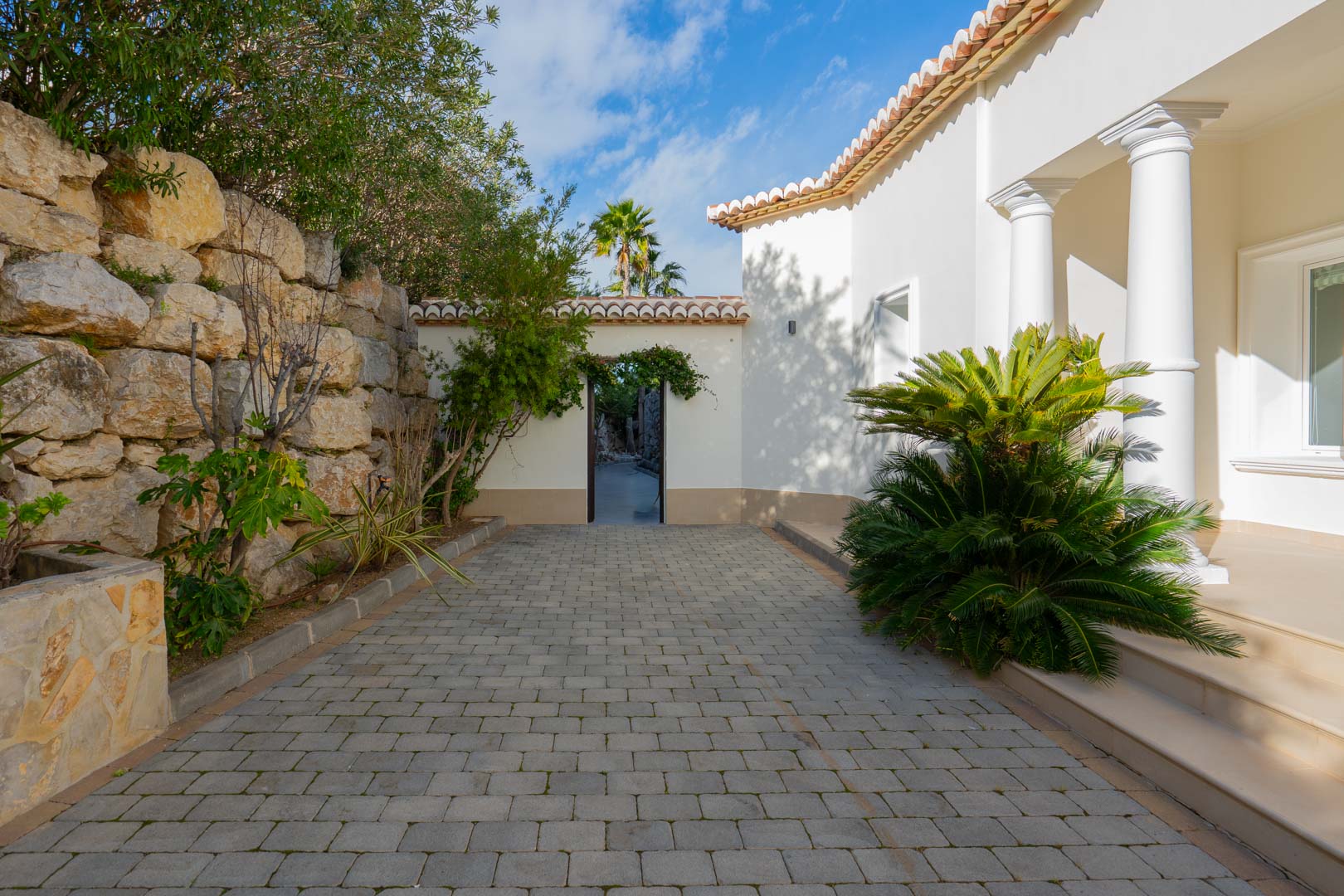 Exterior entrance featuring a grey paved driveway, natural stone retaining wall, and white facade with classical columns and Mediterranean greenery.