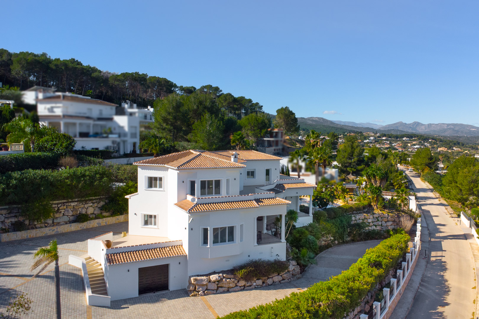 Exterior view of the multi-level villa featuring white facades, terracotta roofing, and stone retaining walls set against a backdrop of Mediterranean mountains.