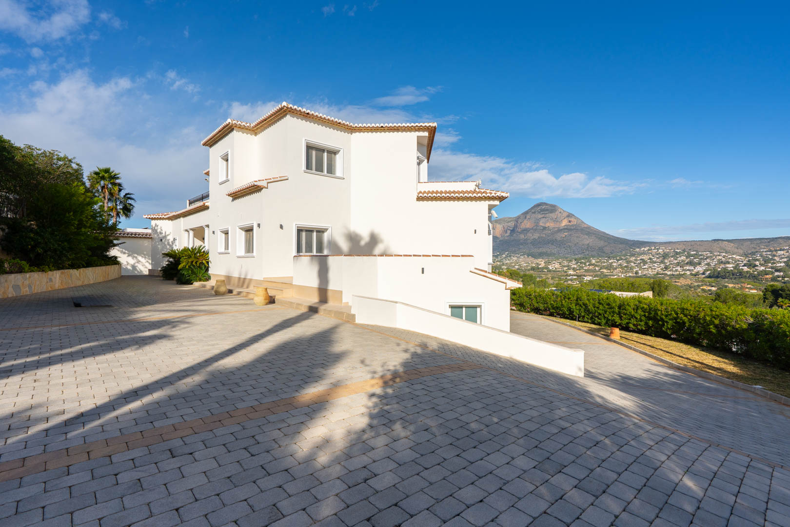 Exterior view of the white-facade villa featuring a large paved driveway and panoramic views of the Montgó mountain under a clear sky.