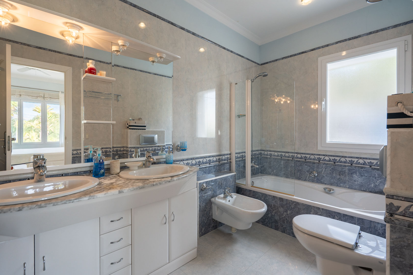 Full bathroom featuring a double vanity with marble countertop, a bathtub with glass partition, and classic blue and grey tiling with decorative borders.
