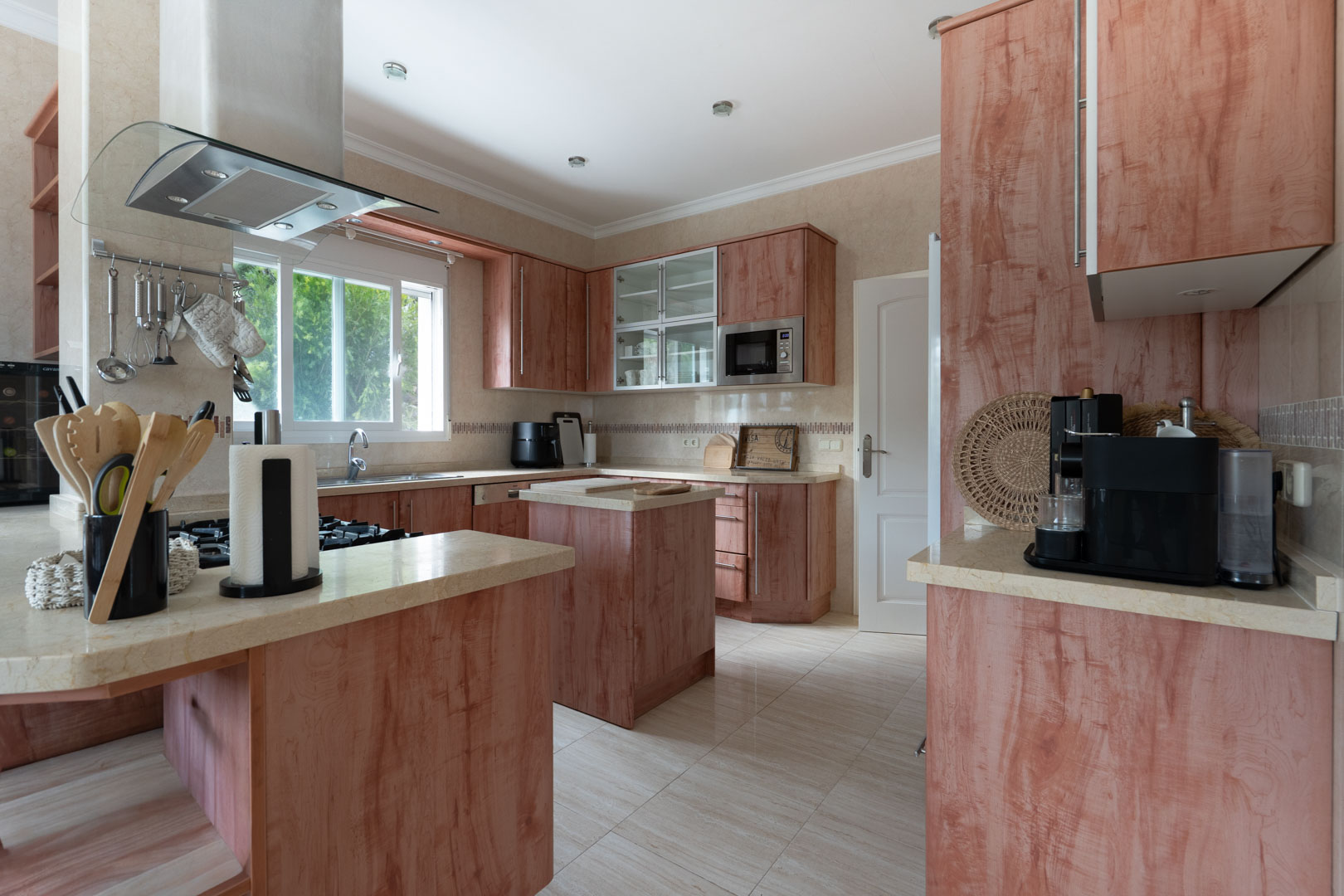 Fully equipped kitchen featuring reddish wood cabinetry, light stone countertops, a central island, and a curved glass extractor hood over the gas cooktop.
