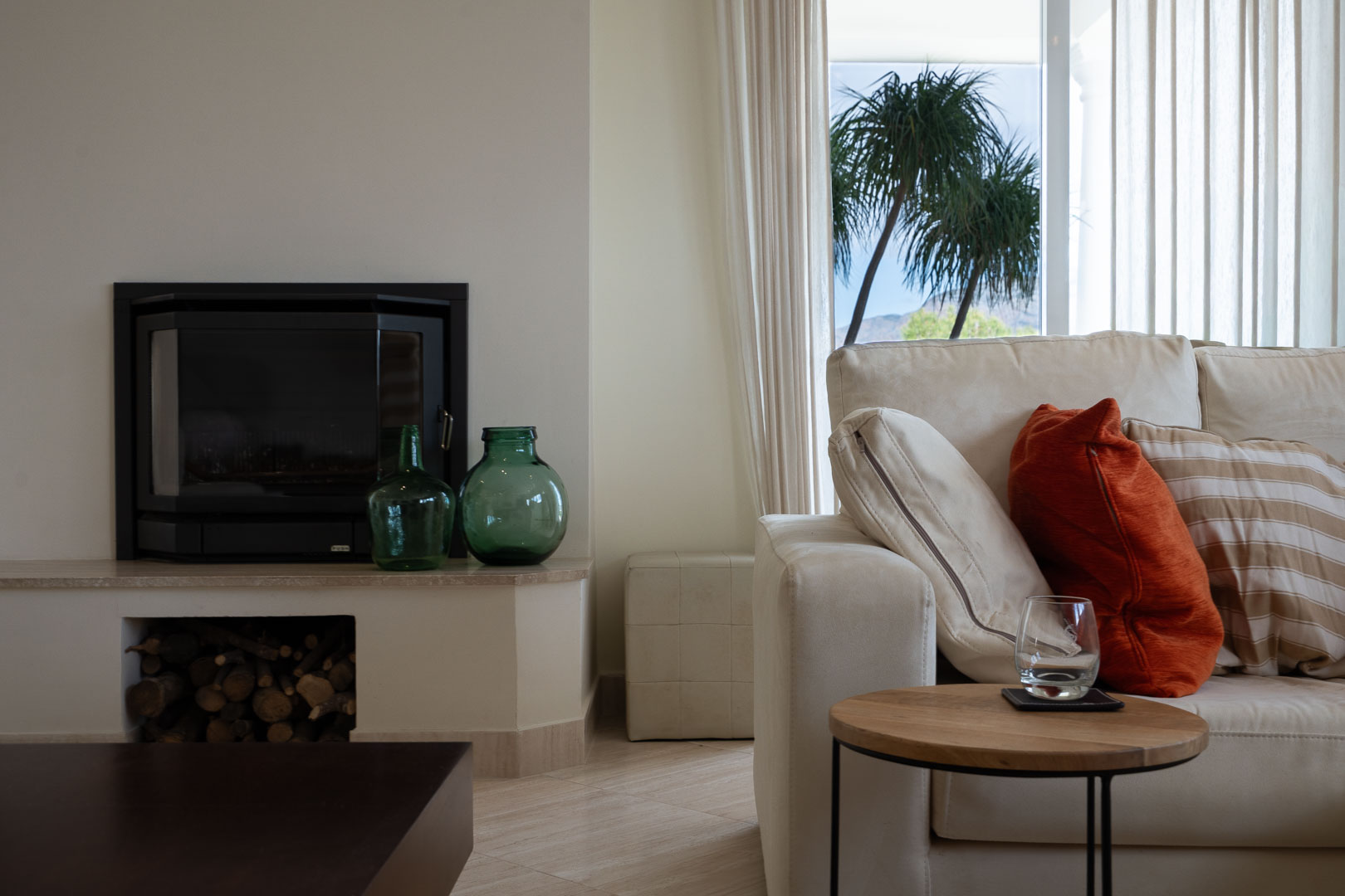 Living area featuring a built-in fireplace with wood storage, neutral-toned sofa, and large windows framing views of palm trees and the Montgó mountain.