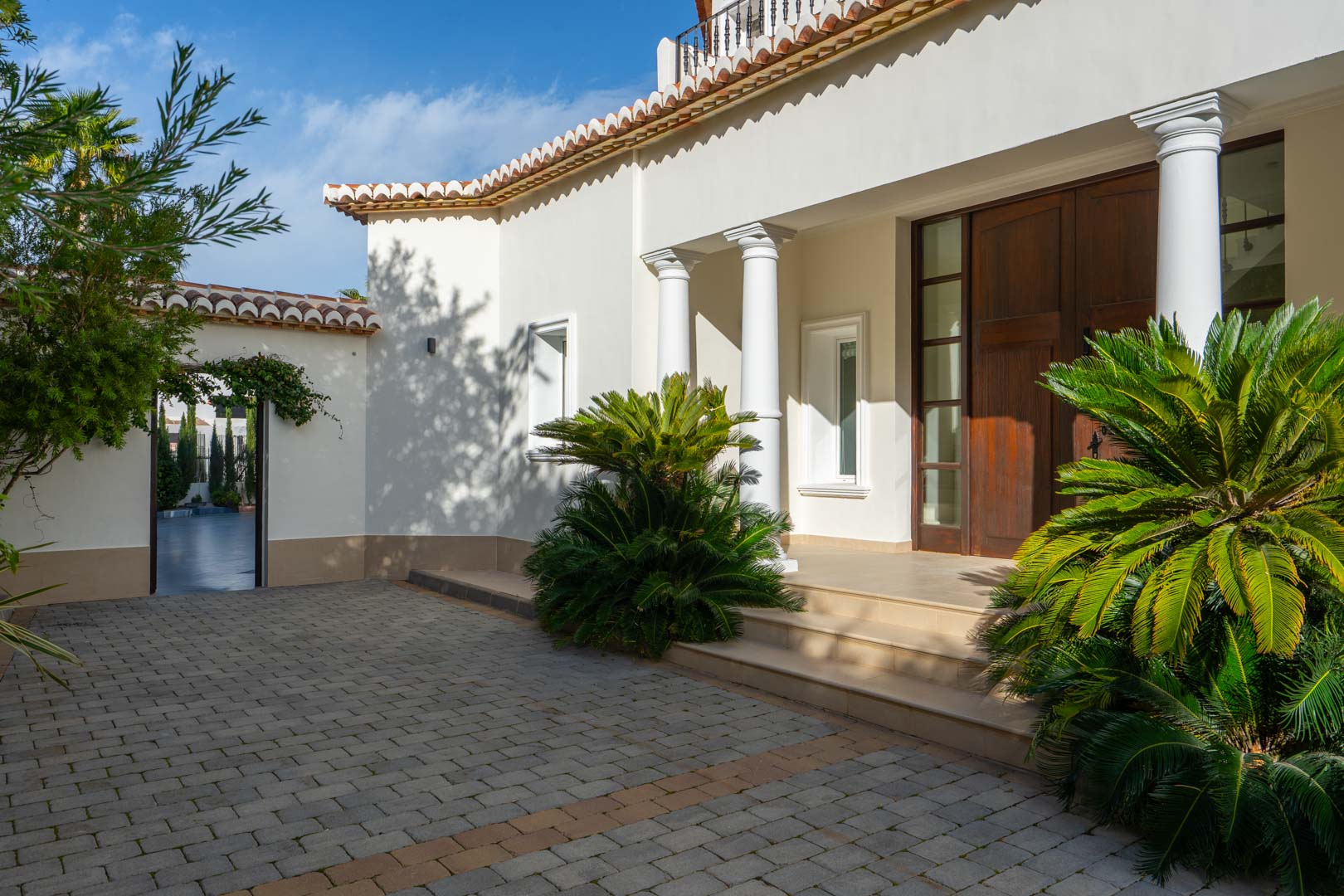 Main entrance featuring a solid wood double door, architectural white columns, and a paved driveway bordered by lush Mediterranean plants.