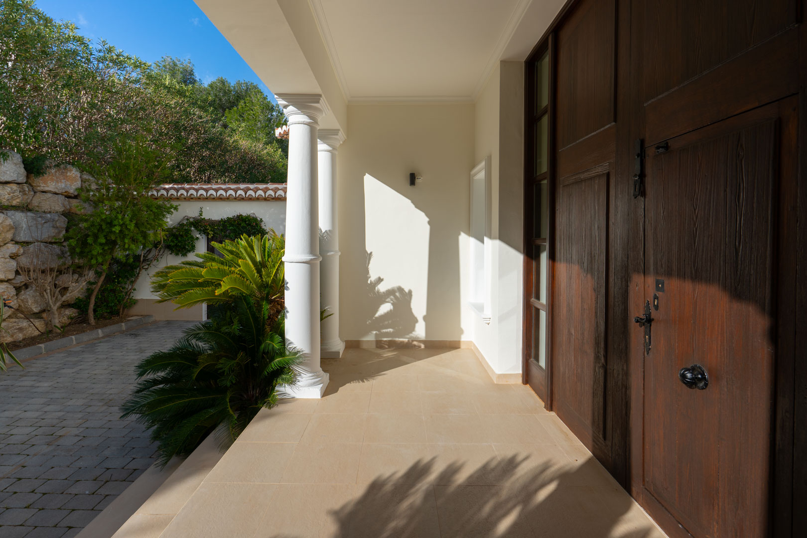 Main entrance featuring a solid wood double door, classical white columns, and a natural stone porch bordered by Mediterranean greenery.