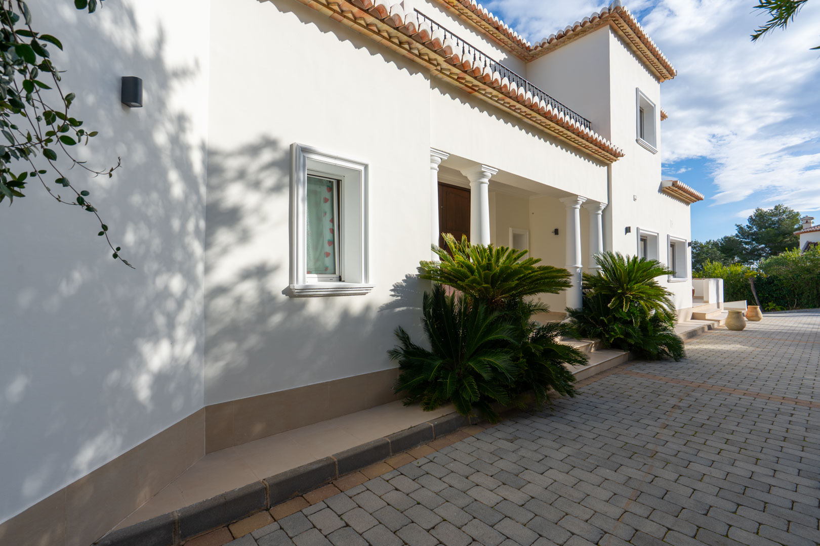 Mediterranean facade featuring white columns, a paved driveway, and architectural details with traditional terracotta roof tiles and lush greenery.