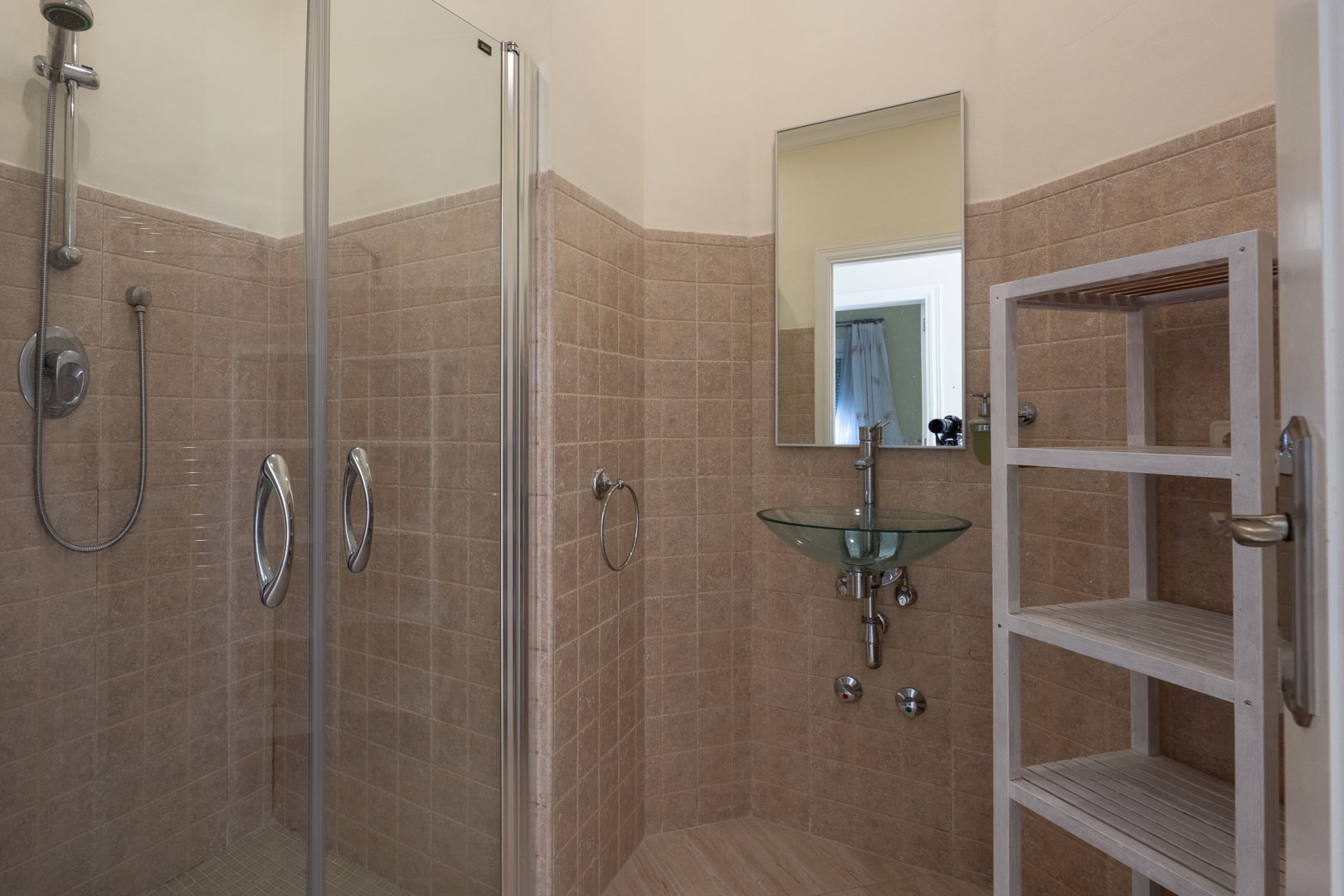 Modern bathroom featuring a glass-enclosed walk-in shower, a unique glass vessel sink, beige tiled walls, and a white wooden slatted storage unit.
