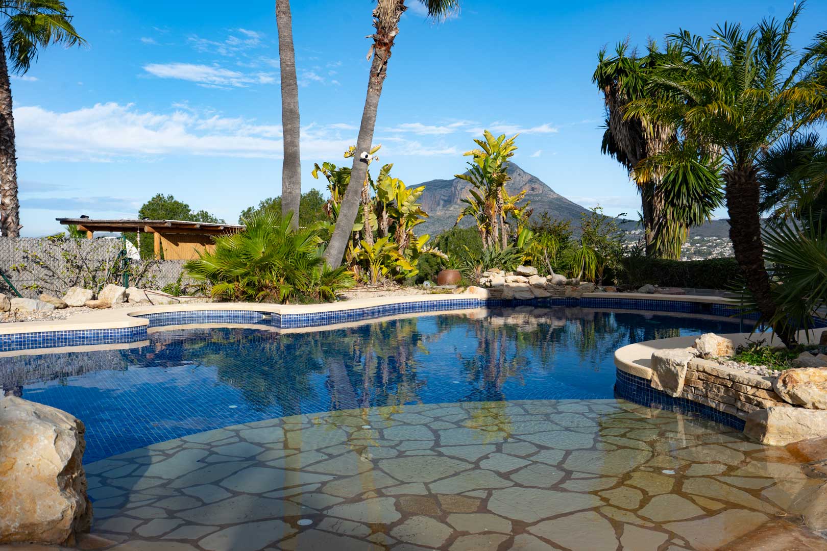 Organic-shaped pool featuring blue mosaic tiles and a natural stone beach entry, set within a tropical garden with clear views of the Montgó mountain.