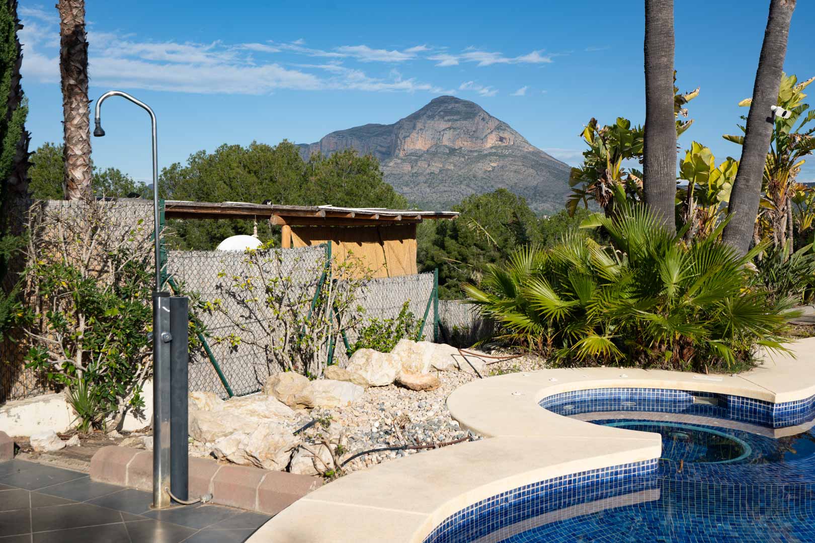 Outdoor pool area featuring a stainless steel shower, lush tropical landscaping, and a clear view of the Montgó mountain.
