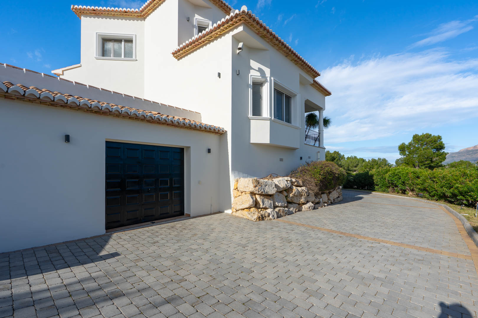 Paved driveway leading to a private garage, featuring white Mediterranean architecture with natural stone retaining walls and traditional terracotta roofing.