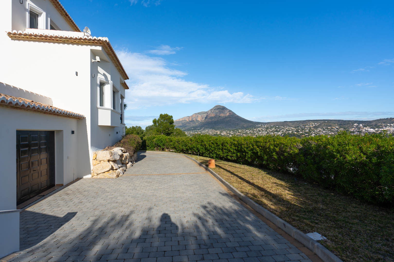 Paved driveway leading to the garage, featuring a natural stone wall and manicured hedges with panoramic views of the Montgó mountain.