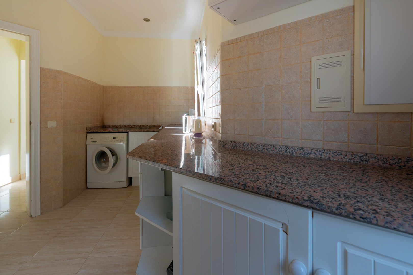 Practical laundry room with granite countertops, white cabinetry, and a washing machine, featuring neutral ceramic tiling and natural light.