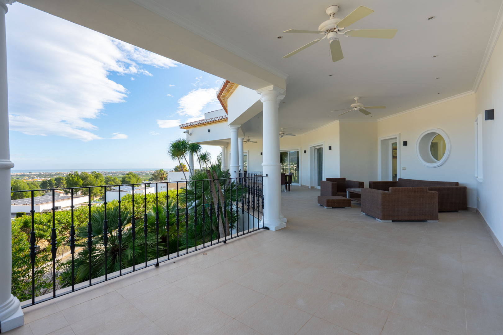 Spacious covered terrace featuring white columns, wrought iron railings, and tiled flooring. Includes an outdoor lounge area with panoramic sea and coastal views.