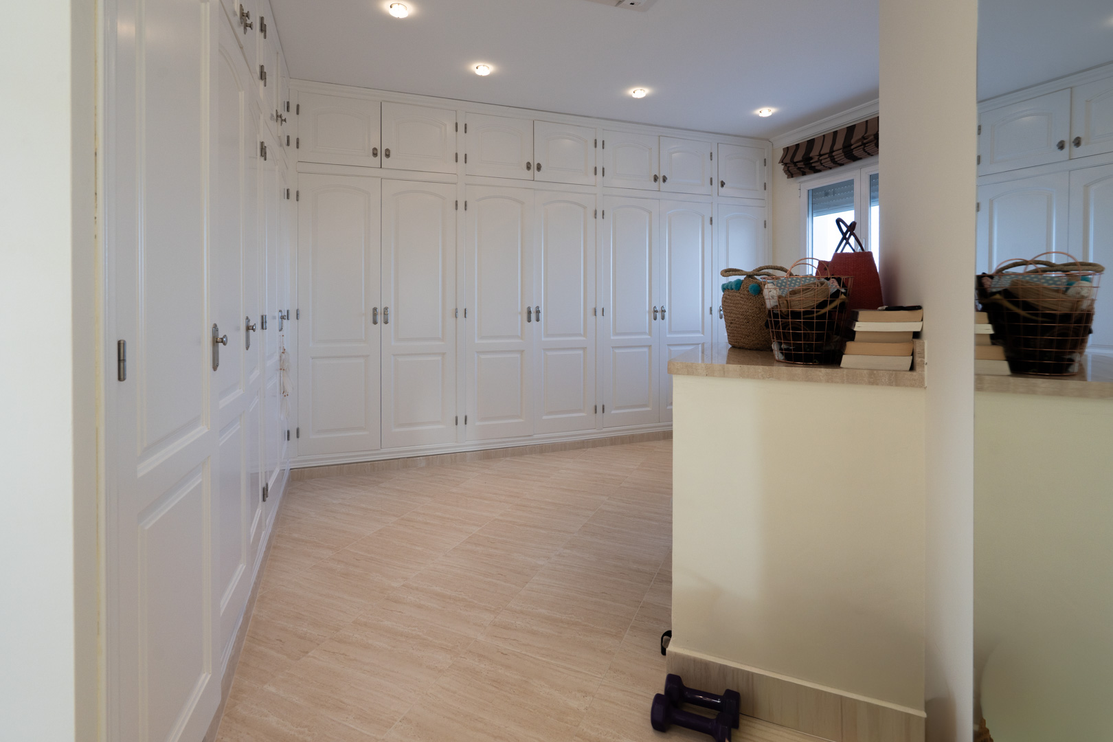 Spacious walk-in closet featuring floor-to-ceiling white paneled wardrobes, recessed ceiling lighting, and neutral-toned tiled flooring.