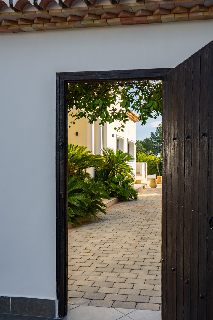 View through a dark wooden door revealing a paved stone pathway, lush Mediterranean greenery, and the elegant white exterior of the villa.