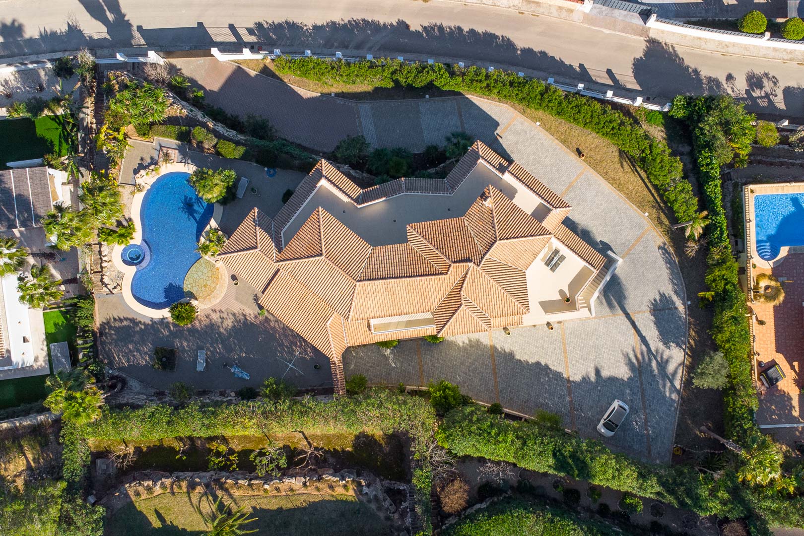 Zenithal aerial view of the property layout, featuring the multi-pitched terracotta roof, a kidney-shaped pool with integrated jacuzzi, and extensive paved parking area.