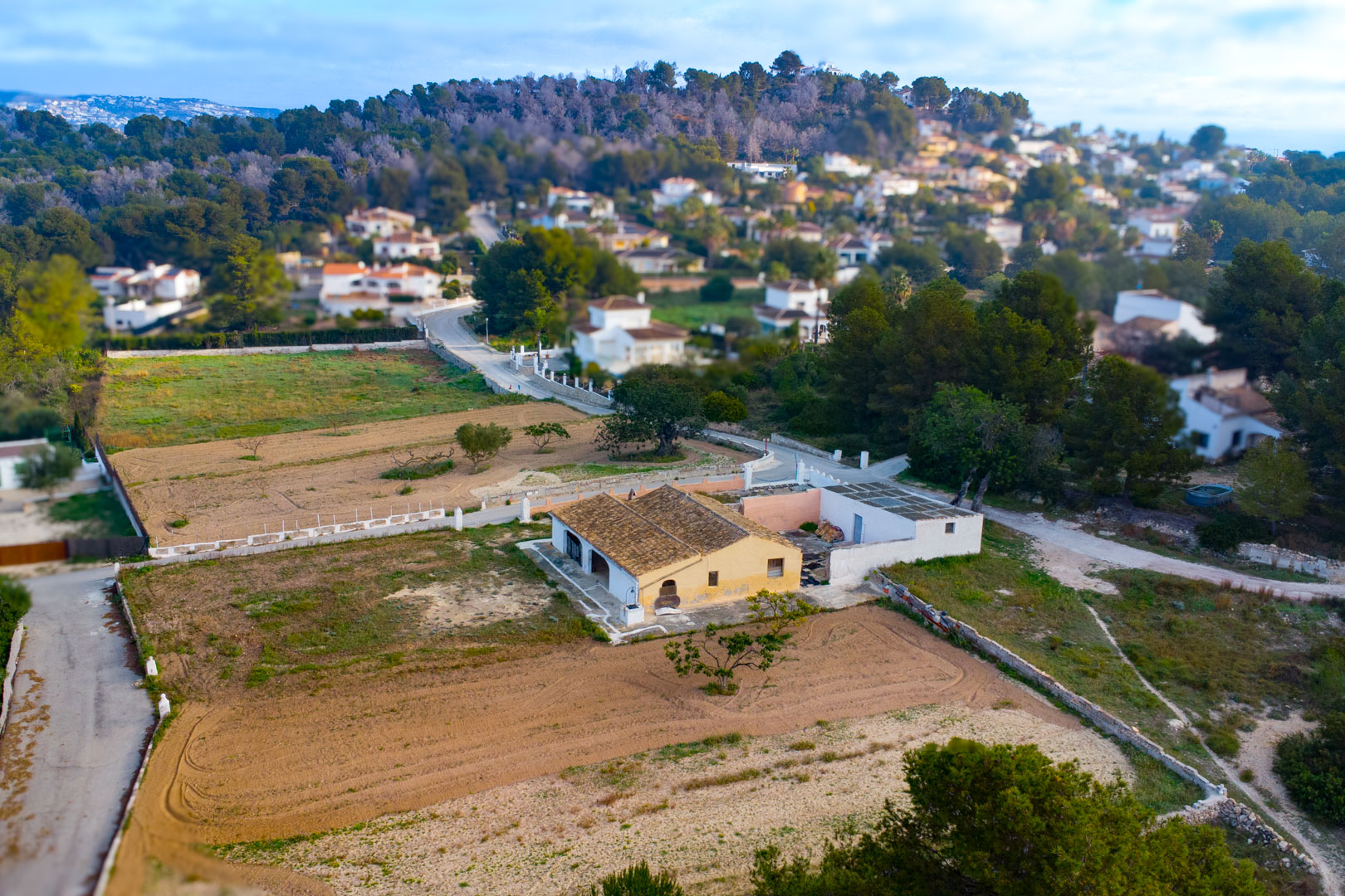 Aerial view of the traditional finca featuring a clay tile roof and ochre facade, set on a large rustic plot surrounded by Mediterranean greenery and villas.