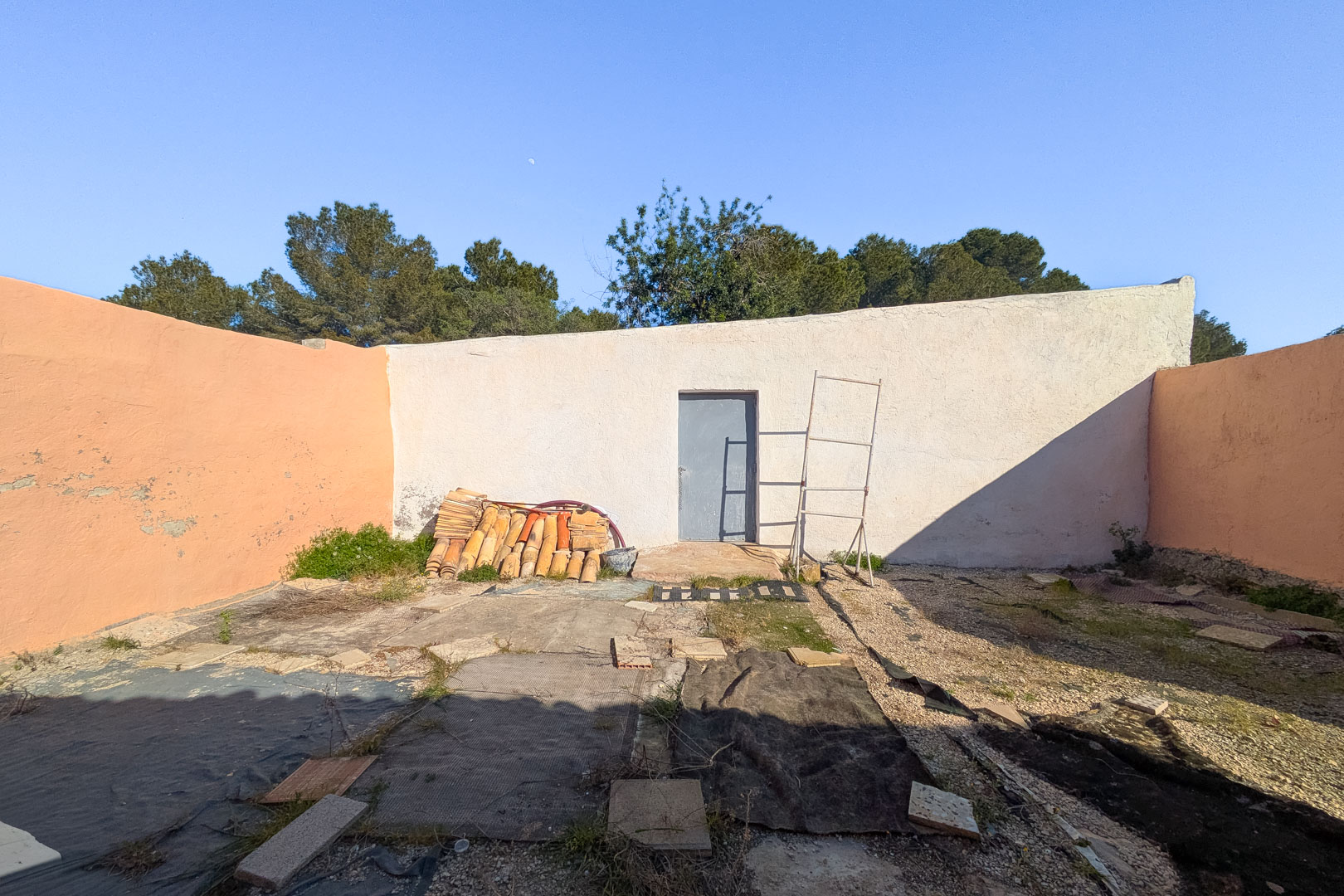 Enclosed outdoor courtyard featuring high perimeter walls, a central access door, and stacked traditional roof tiles on a rustic ground surface.
