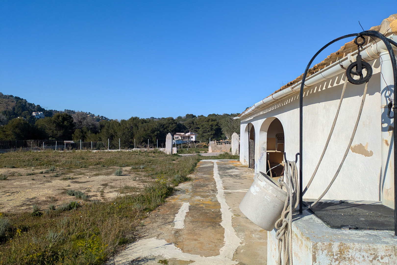 Exterior of the finca featuring a traditional stone well with pulley, adjacent to the white arched facade and a large flat rustic plot with mountain views.
