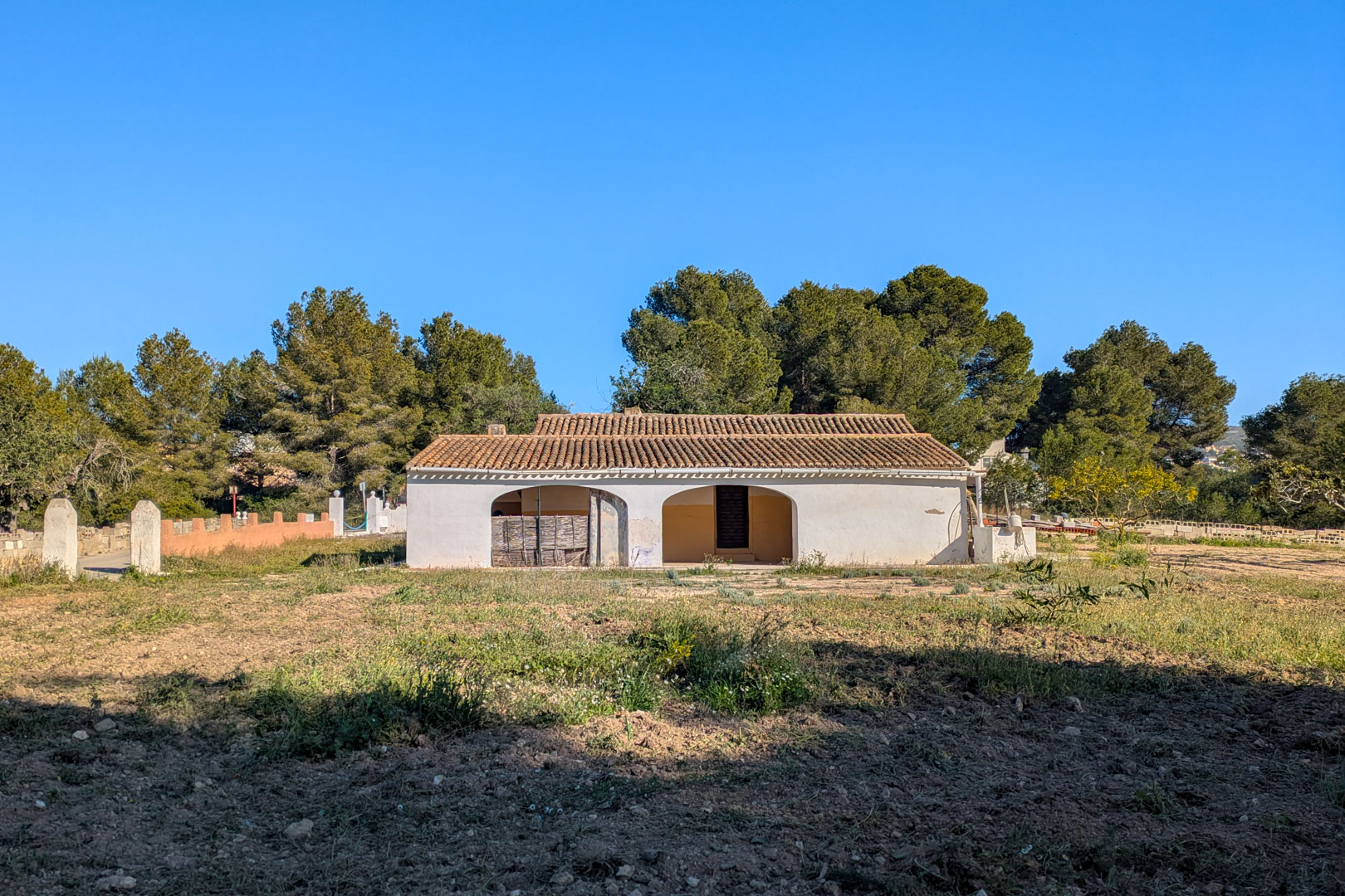 Exterior view of the traditional finca featuring a white facade, arched porch, and terracotta tile roof, set on a rustic plot surrounded by pine trees.