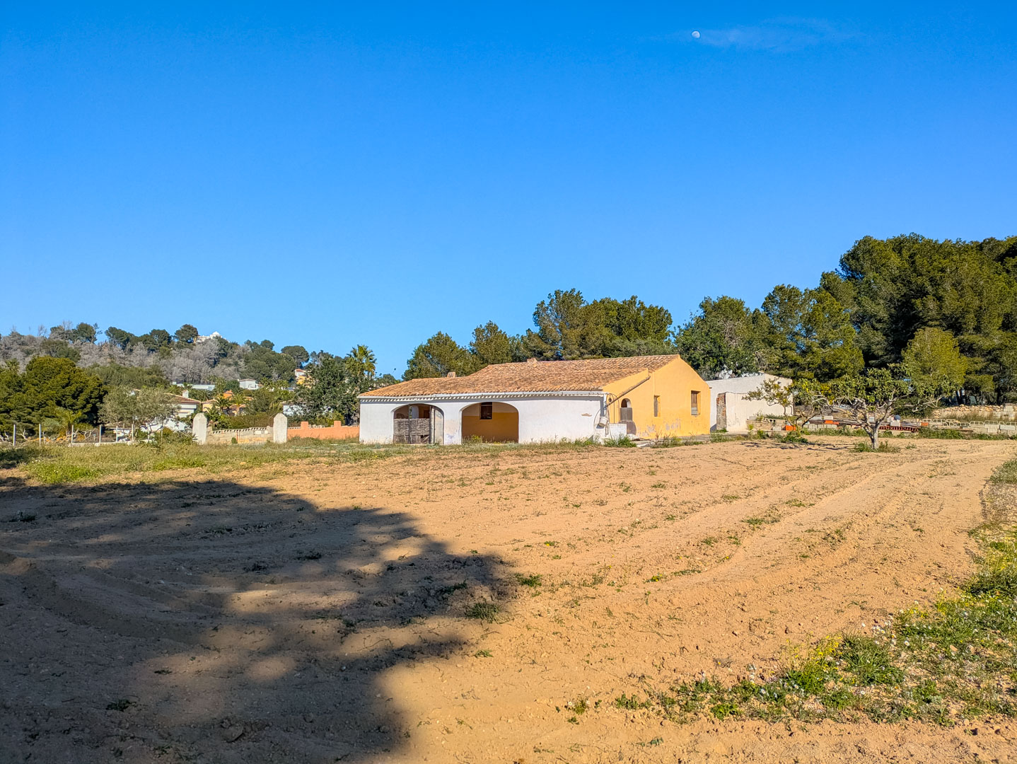 Exterior view of the traditional finca featuring arched porch entries, a terracotta tile roof, and a vast flat rustic plot bordered by Mediterranean pines.