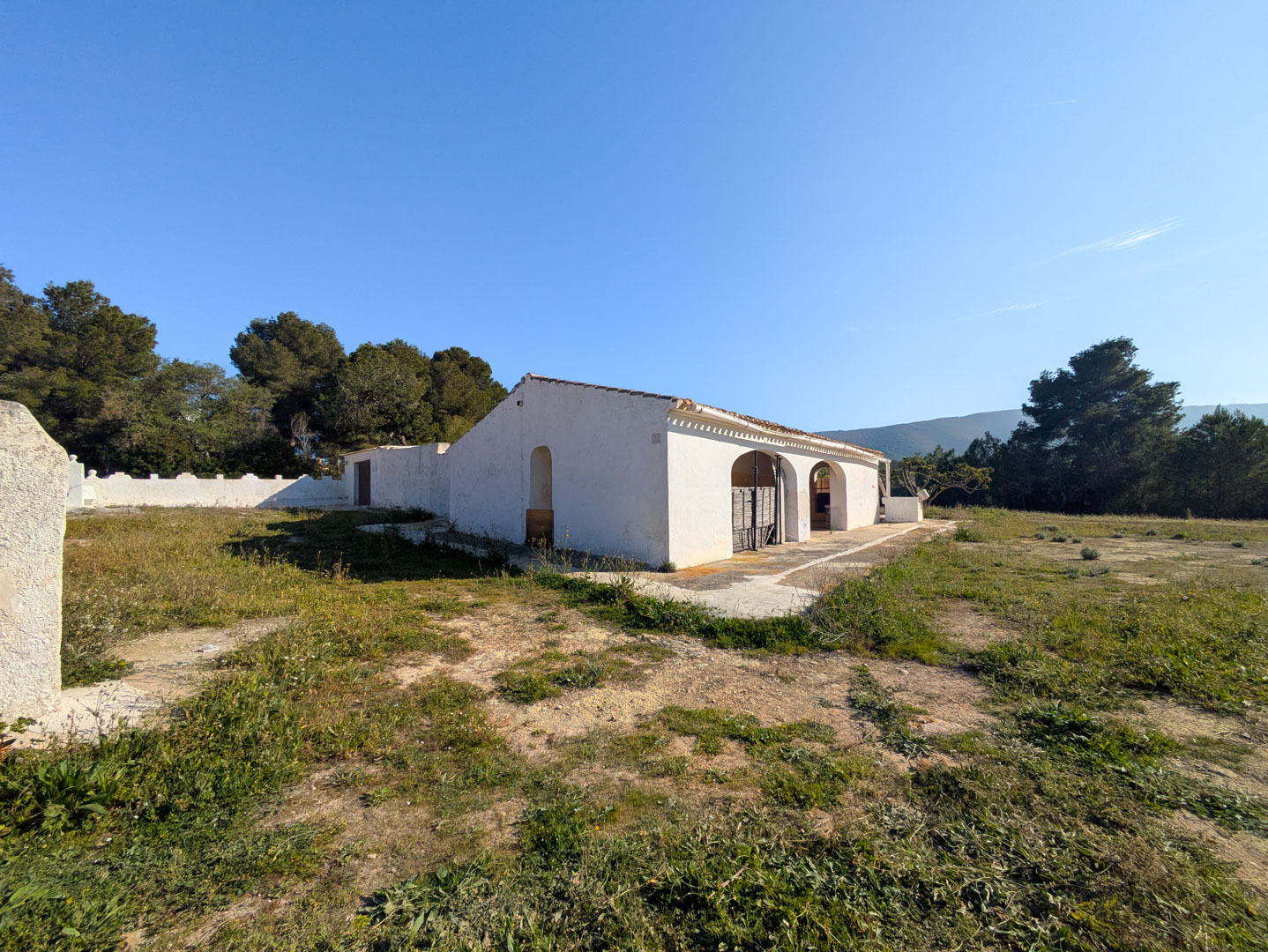 Exterior view of the traditional finca with white-washed walls and arched naya, set on a flat rustic plot surrounded by Mediterranean pine trees.