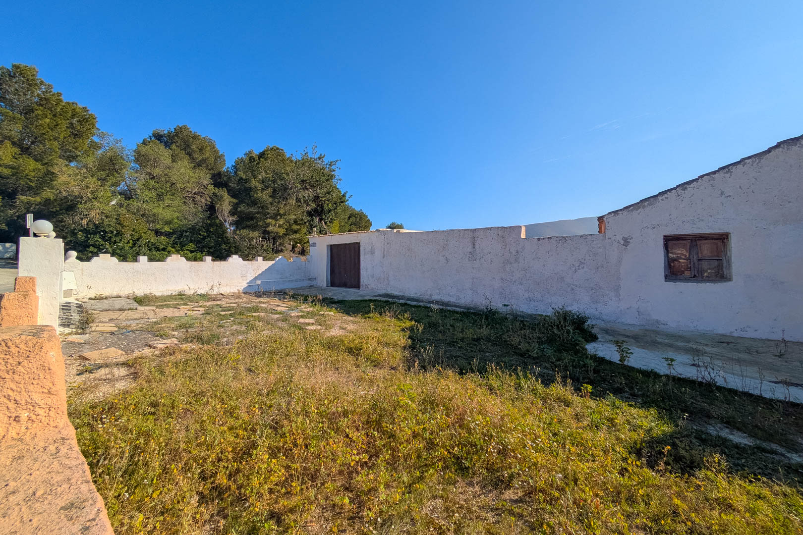 Exterior view of the whitewashed facade featuring original wooden joinery and a walled rustic courtyard surrounded by Mediterranean pines and natural vegetation.