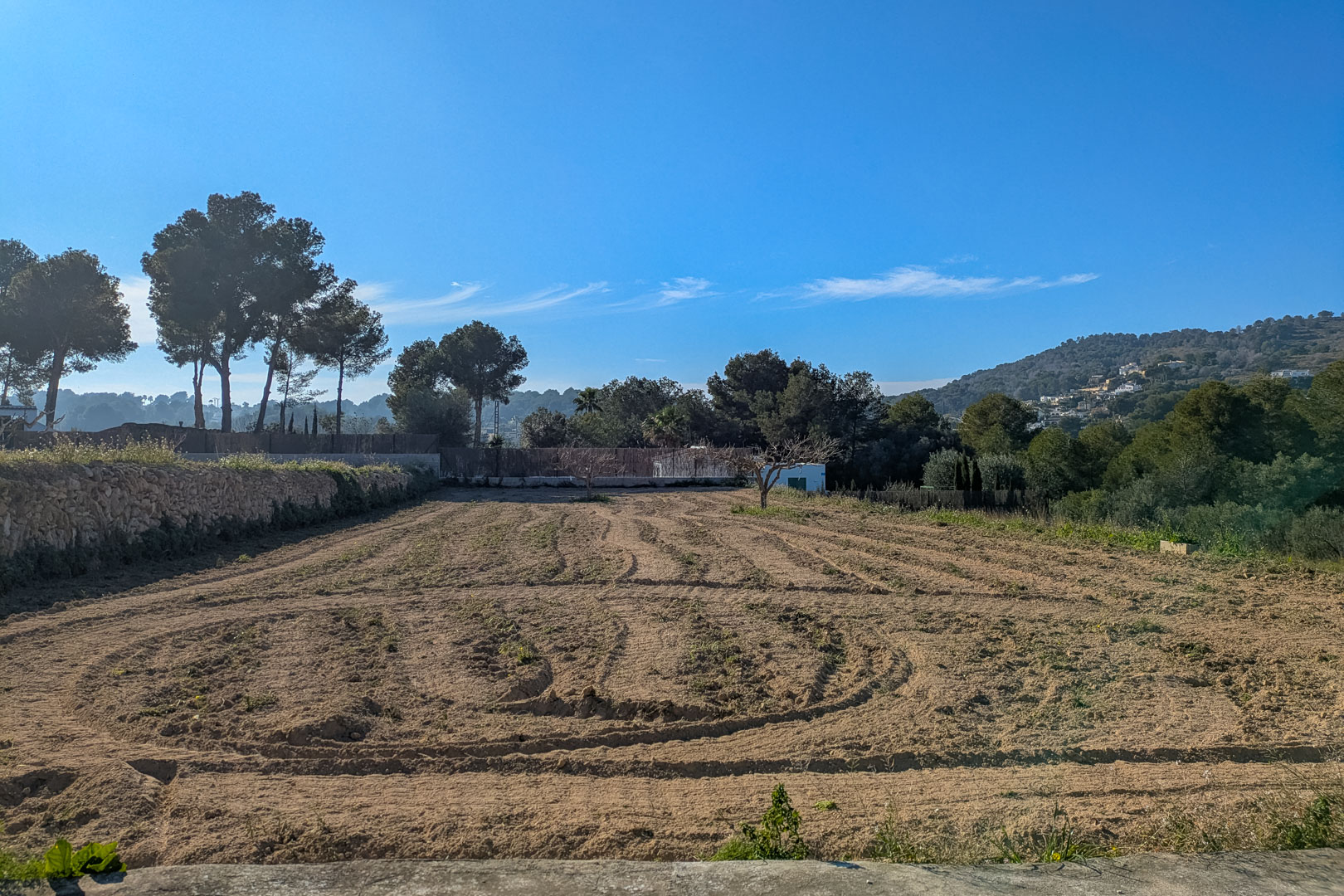 Flat, tilled rustic plot bordered by a traditional stone wall, featuring open views of the surrounding hills and mature pine trees under a clear blue sky.