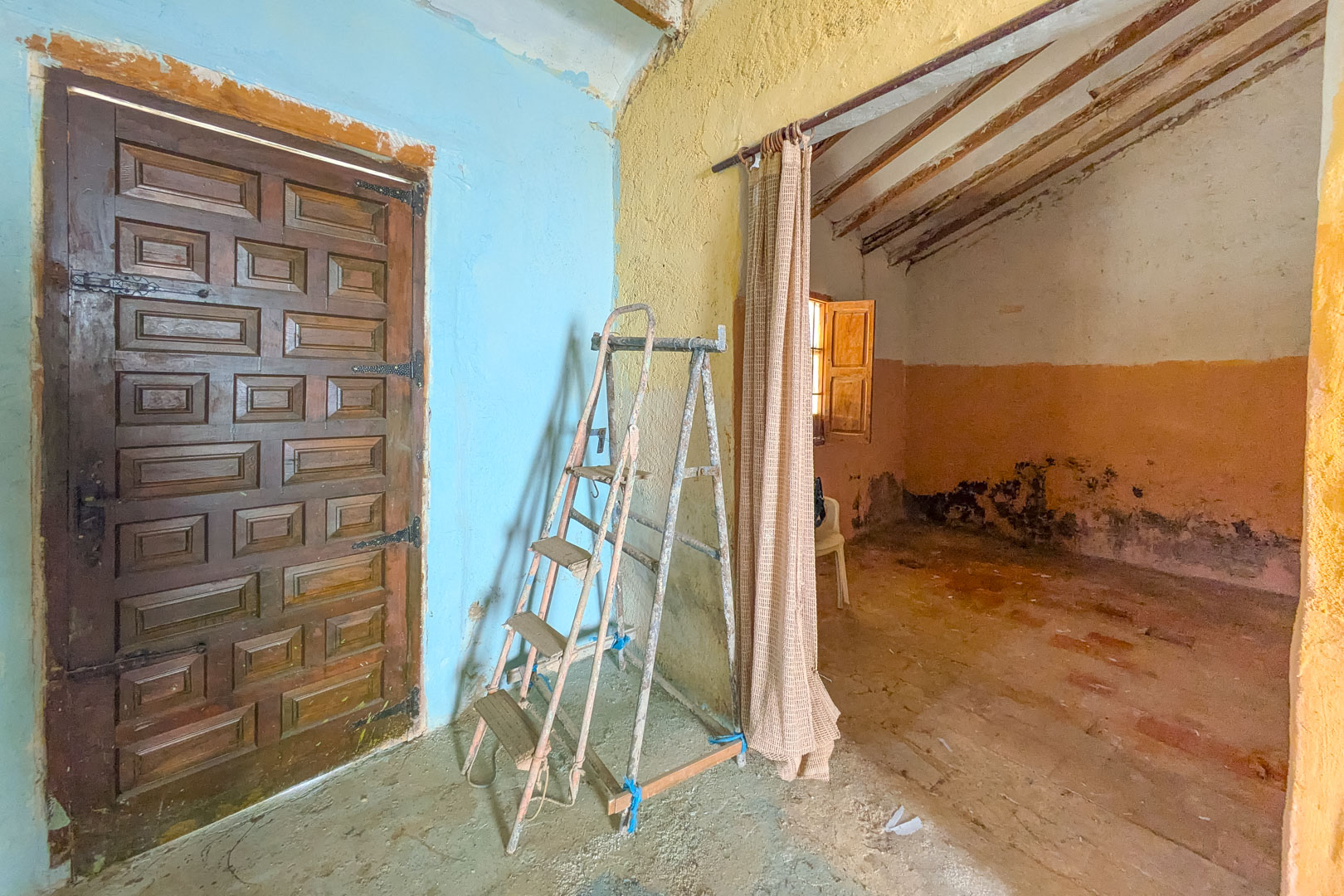 Rustic interior featuring a traditional paneled wood door, textured walls in blue and yellow, and original ceiling beams visible in the adjacent room.