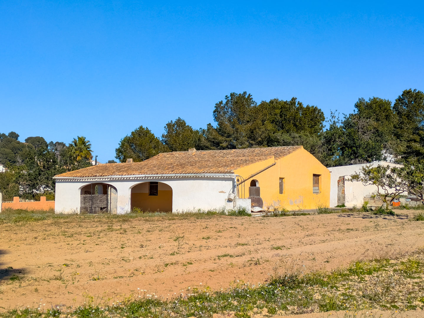 Traditional finca facade featuring arched porches and a clay tile roof, set on a vast rustic plot surrounded by mature pine trees and nature.