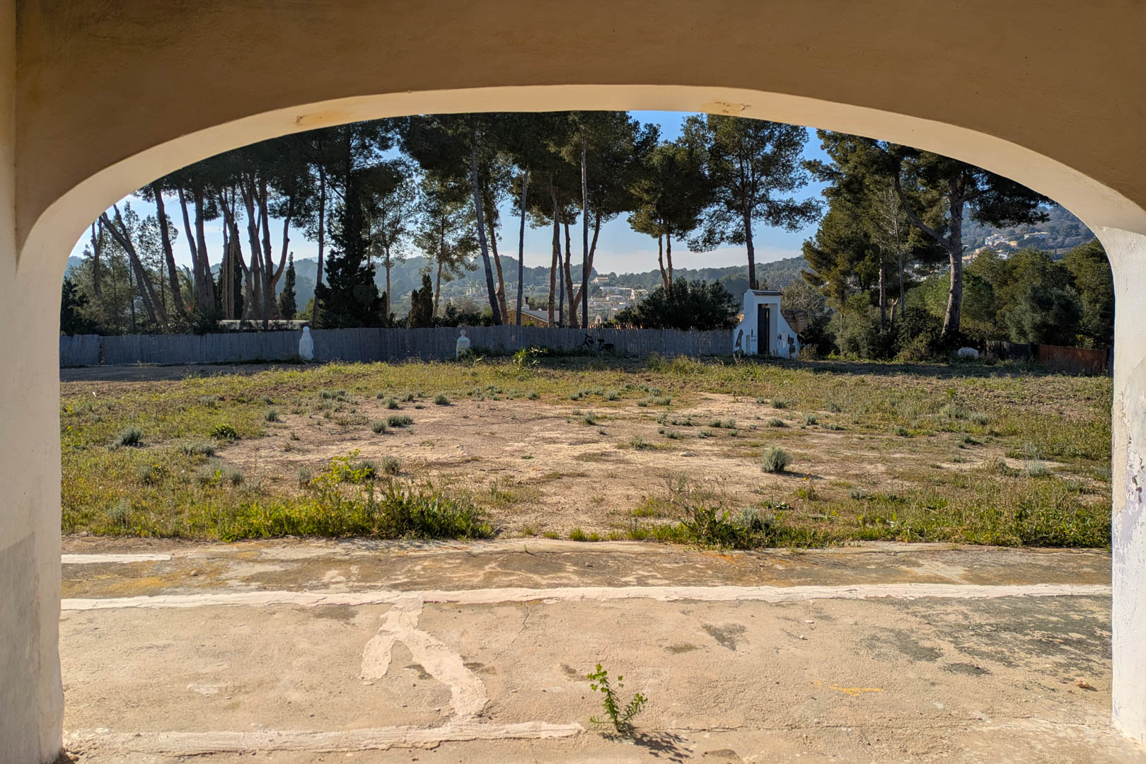 View through a traditional arched porch overlooking the expansive flat plot, bordered by mature pine trees and natural landscape.