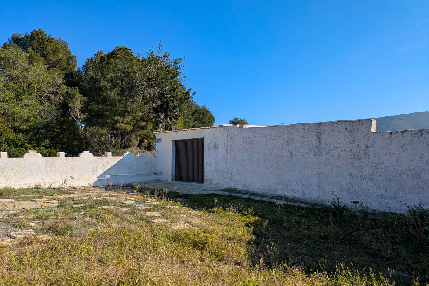 White-washed perimeter wall featuring a dark access door, set within a rustic plot surrounded by Mediterranean pines and natural vegetation.