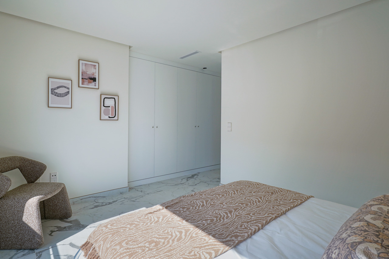 Bright bedroom featuring floor-to-ceiling white wardrobes, marble-effect flooring, and a modern armchair accented by minimalist wall art.