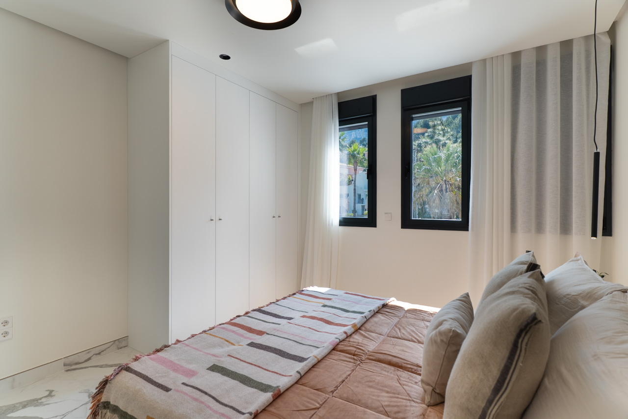 Bright bedroom featuring floor-to-ceiling white wardrobes, marble-effect flooring, and windows overlooking the Mediterranean landscape.