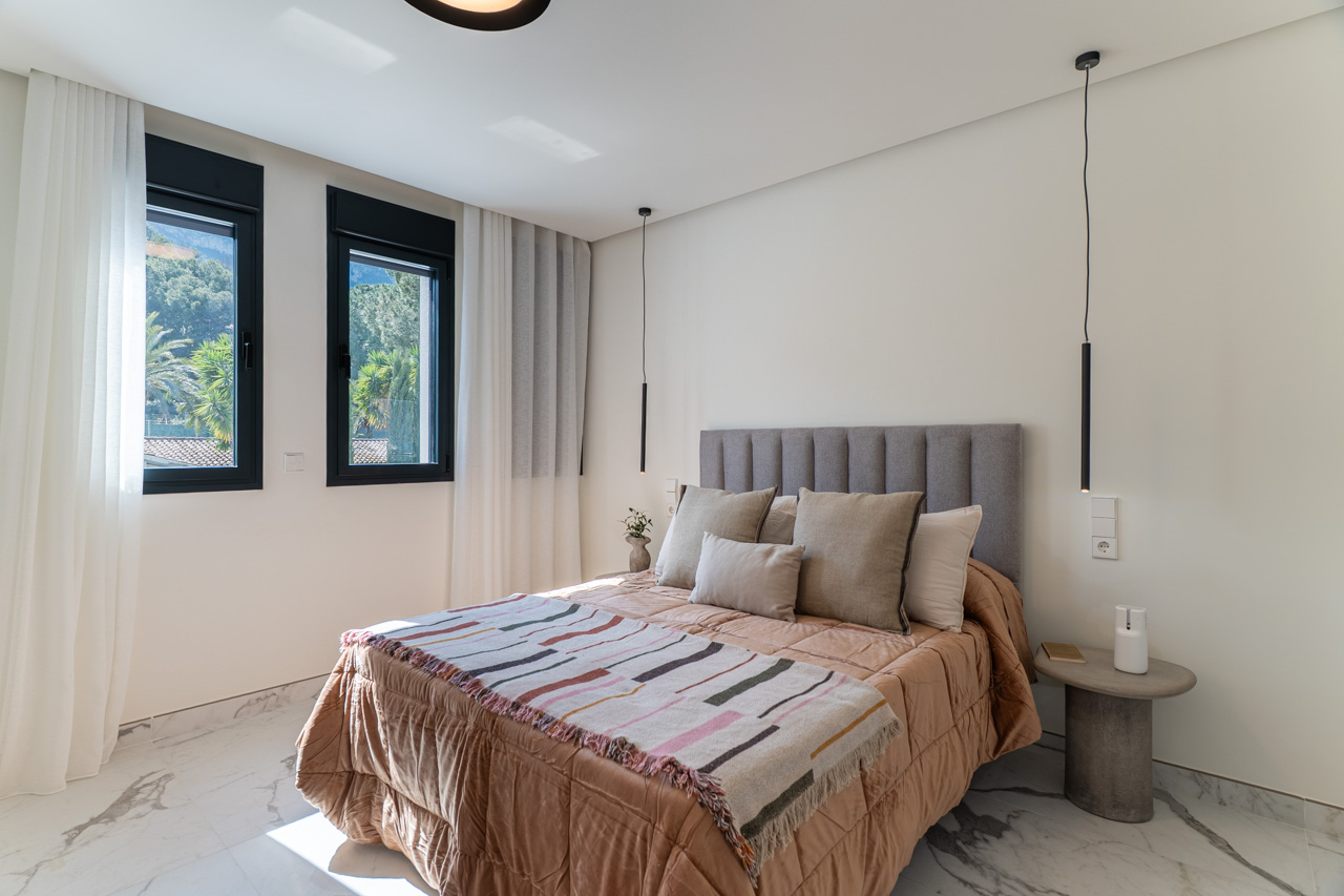Double bedroom featuring marble-effect porcelain flooring, a grey upholstered headboard, and black-framed windows providing natural light and views of the greenery.