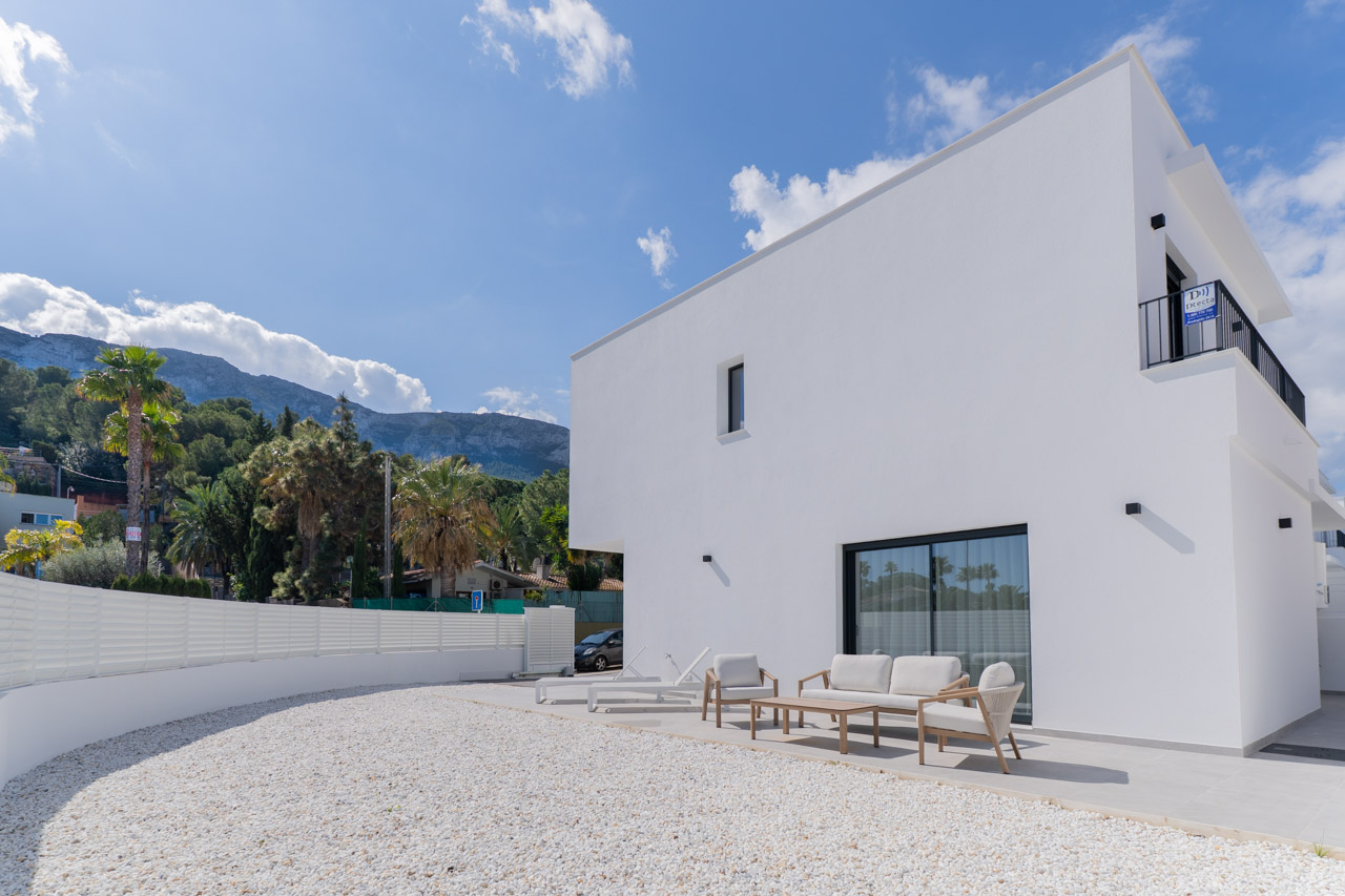 Minimalist white villa exterior featuring a tiled terrace with lounge furniture, a white gravel garden, and views of the Montgó mountain range.