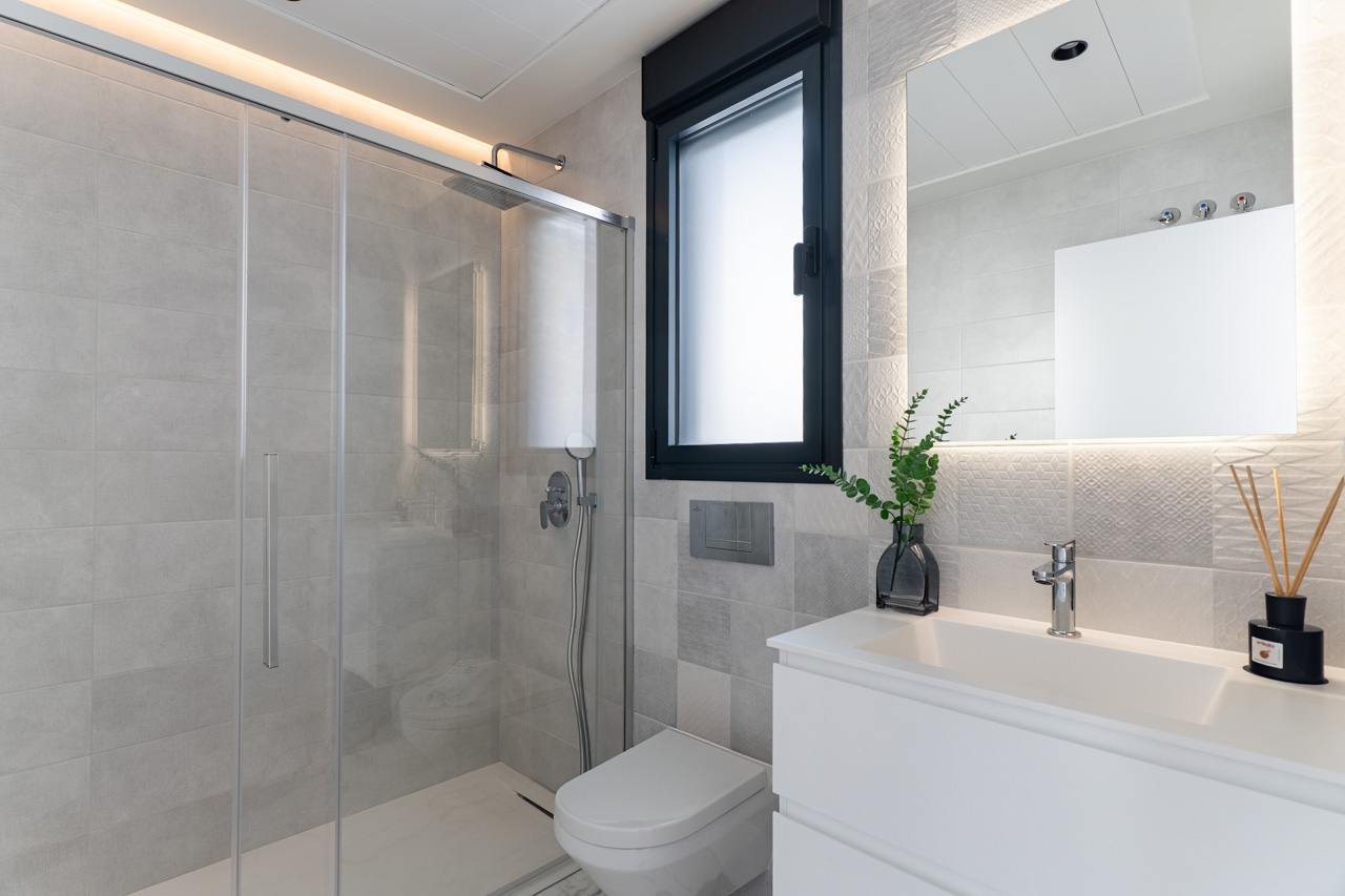 Modern bathroom featuring a walk-in glass shower, textured grey wall tiles, a white vanity unit, and a large backlit mirror with integrated LED lighting.