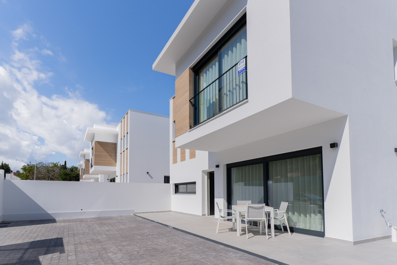 Modern exterior featuring white facades with wood-effect accents, a paved private terrace, and outdoor dining furniture set against large sliding glass doors.