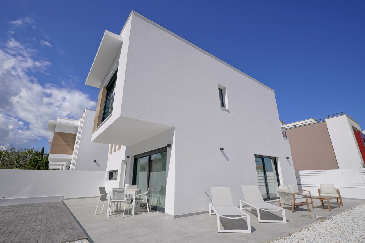 Modern exterior featuring white geometric volumes, a tiled terrace equipped with outdoor seating and loungers, and a private paved parking area.