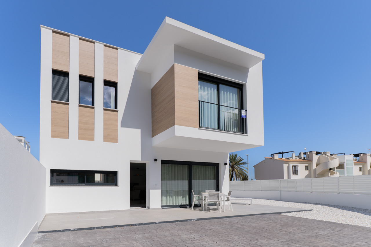 Modern exterior featuring wood-effect cladding, a paved terrace with outdoor dining furniture, and large black-framed windows under a clear sky.